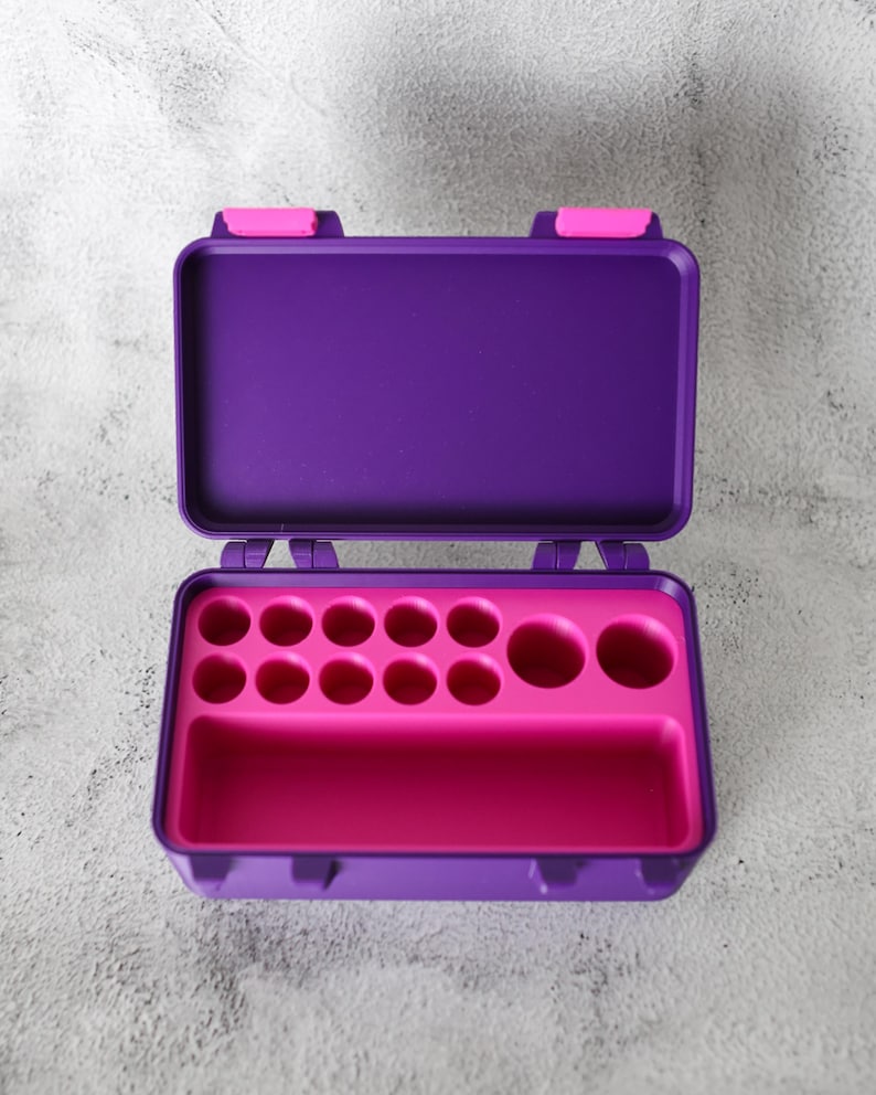 May include: Open purple and pink plastic storage case. The case has a hinged lid with pink clasps. Inside, a pink tray features various circular compartments and a rectangular section, designed for organizing small items.
