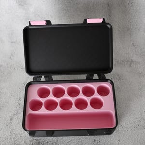 May include: Open black carrying case with pink interior. The case has a rectangular shape with a hinged lid and pink latches. The interior features circular compartments and a rectangular slot.