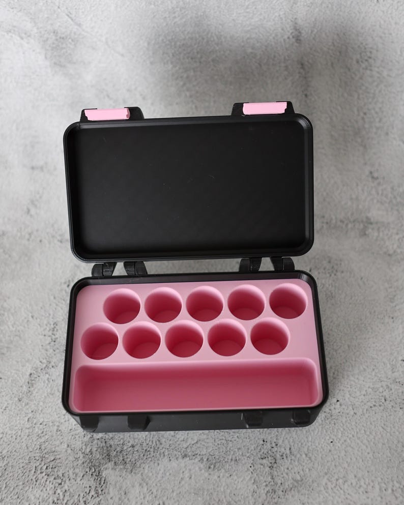 May include: Open black carrying case with pink interior. The case has a rectangular shape with a hinged lid and pink latches. The interior features circular compartments and a rectangular slot.