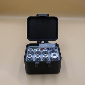 May include: A black rectangular storage case with an open hinged lid. The interior features a grey insert holding six small glass vials with silver caps, one larger glass vial with a silver cap, and a stack of small rectangular cards.