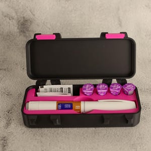 May include: Black case with a pink interior, containing a white medical pen, several purple medication vials, and a small packet of papers. The case has pink clasps and hinges.
