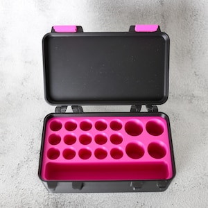May include: Open black case with a bright pink interior. The case has a rectangular shape with a hinged lid and a series of circular cutouts inside. The lid has two pink accents.