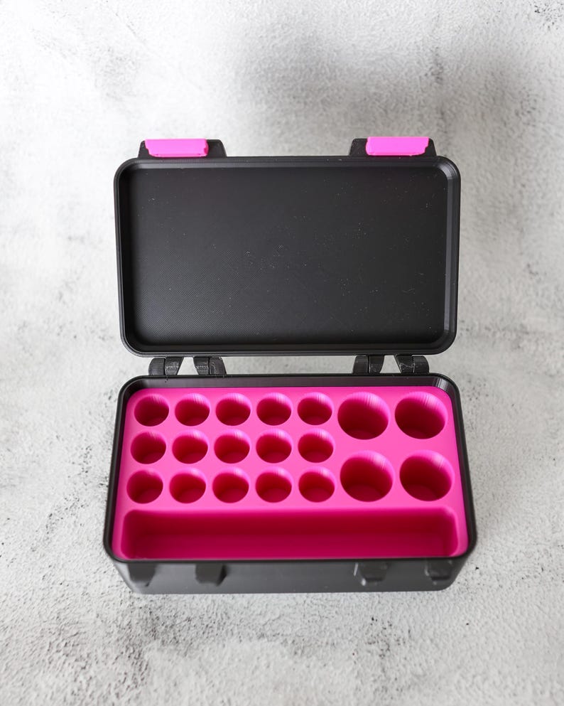 May include: Open black case with a bright pink interior. The case has a rectangular shape with a hinged lid and a series of circular cutouts inside. The lid has two pink accents.