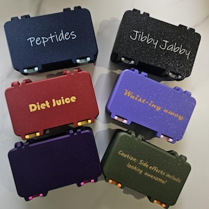 May include: Six small, colorful cases with hinged lids. Each case has text on the top: "Peptides", "Jibby Jabby", "Diet Juice", "Waist-ing away", and "Caution: Side effects include looking awesome!". The cases are navy, black, red, purple, dark purple, and olive green.