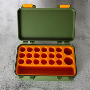 May include: Open green plastic storage box with orange interior. The box has a rectangular shape with a hinged lid and orange latches. The interior features multiple circular compartments and a rectangular slot.
