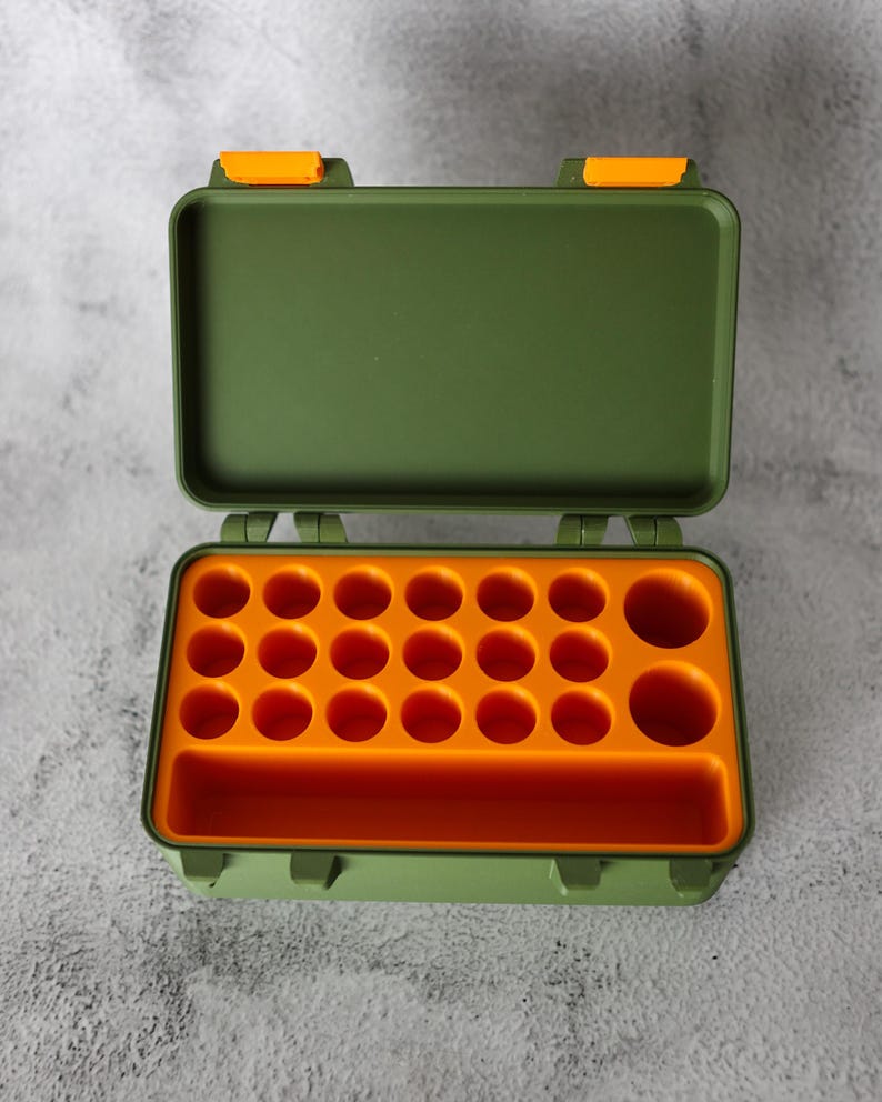 May include: Open green plastic storage box with orange interior. The box has a rectangular shape with a hinged lid and orange latches. The interior features multiple circular compartments and a rectangular slot.