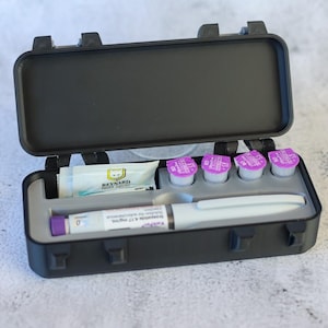 May include: A black plastic case with a hinged lid, containing a medical pen, four small vials with purple tops, and a small white packet with the text "REYNAUD" printed on it.