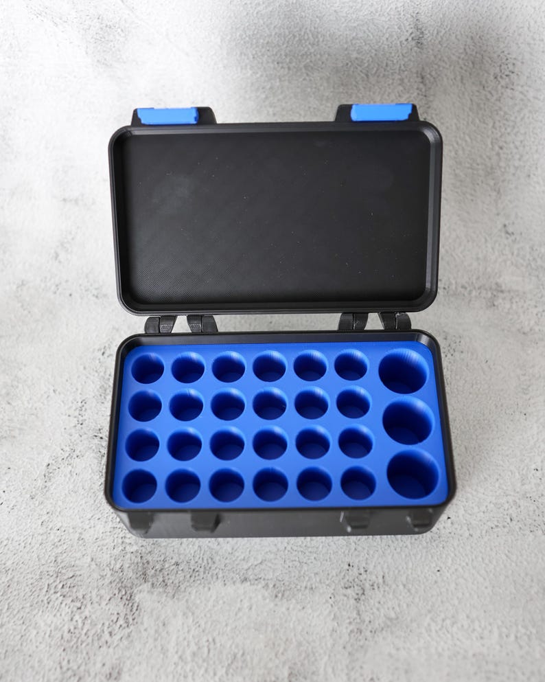 May include: An open black plastic case with a blue interior. The case has a rectangular shape with a hinged lid and blue clasps. The interior has multiple circular compartments of varying sizes.