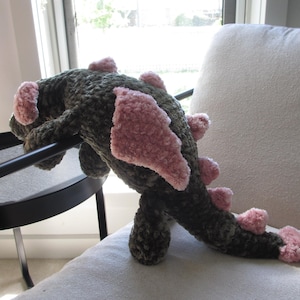 May include: A handmade plush dragon toy in shades of green and pink. The dragon has a textured, fuzzy body with pink accents on its wings and back. It is resting on a white chair and a black table.