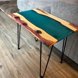 May include: A teal epoxy resin river table with a natural wood base and black hairpin legs. The table is sitting on a wood floor.