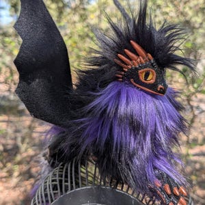 May include: A handcrafted fantasy dragon sculpture with a black body, purple fur, and copper-colored accents. The dragon has large black wings and is perched on a metal object. The dragon's head features yellow eyes and a detailed, textured design.