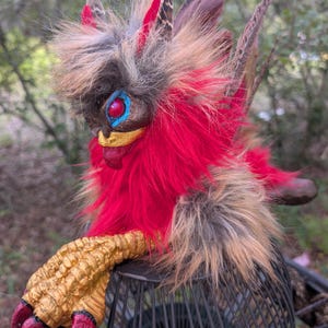 May include: A fantasy creature figurine featuring vibrant red and tan faux fur, a gold-painted clawed hand with red-tipped talons, and a face with bright red eyes, blue accents, and a gold beak. The creature has long brown feathers extending from its back.