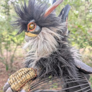 May include: A fantasy creature figurine with black and grey faux fur, a gold-painted textured clawed hand, and a sharp beak. The creature features a prominent crest of black hair, a single visible amber-colored eye, and pointed ears.