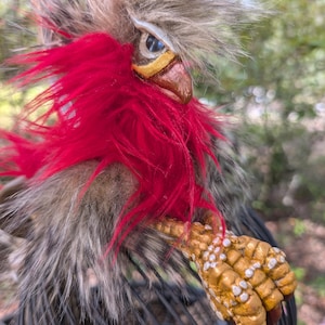 May include: A handcrafted bird figurine featuring long, shaggy grey faux fur, a vibrant red feathered chest, and a prominent crest. The bird has a brown beak, a single visible blue eye, and textured, golden-yellow talons with white accents.