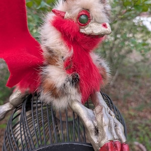 May include: A handmade fantasy creature with red wings and accents, perched on a black wire cage. The creature has a mix of brown, white, and red fur, with gold-painted details on its face and feet. Its large green eyes give it a whimsical appearance.