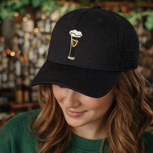 May include: Black baseball cap with a mesh back and a beer glass graphic. The graphic is yellow and white, with a black outline. The cap is worn by a person with long, wavy brown hair.