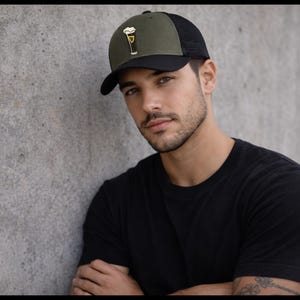 May include: A black and olive green baseball cap with a mesh back. The cap features an embroidered design of a pint of beer. The brim is black, and the crown is olive green with a black mesh back.