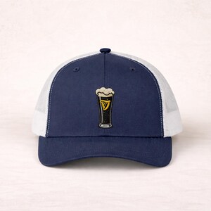 May include: Navy blue trucker hat with a white mesh back and a curved brim. The hat features a patch of a pint of stout with a foamy head and a gold harp logo. A stylish accessory for casual wear.
