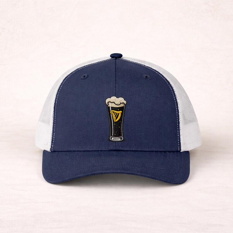 May include: Navy blue trucker hat with a white mesh back and a curved brim. The hat features a patch of a pint of stout with a foamy head and a gold harp logo. A stylish accessory for casual wear.