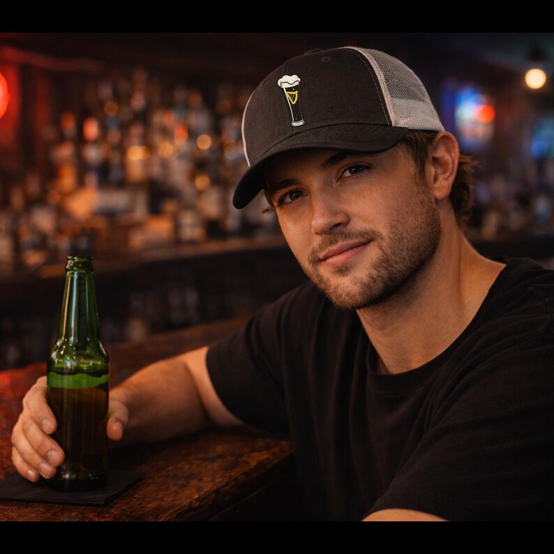 May include: Black and white trucker hat with a beer glass graphic. The hat has a black front panel and brim, a white mesh back, and a beer glass design on the front. A green beer bottle is on the table.