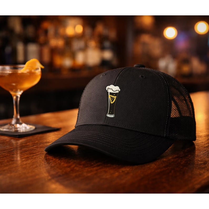 May include: Black baseball cap with a mesh back and a curved brim. The cap features an embroidered design of a pint of beer with a white foamy head and a gold harp symbol. The cap is sitting on a wooden bar.