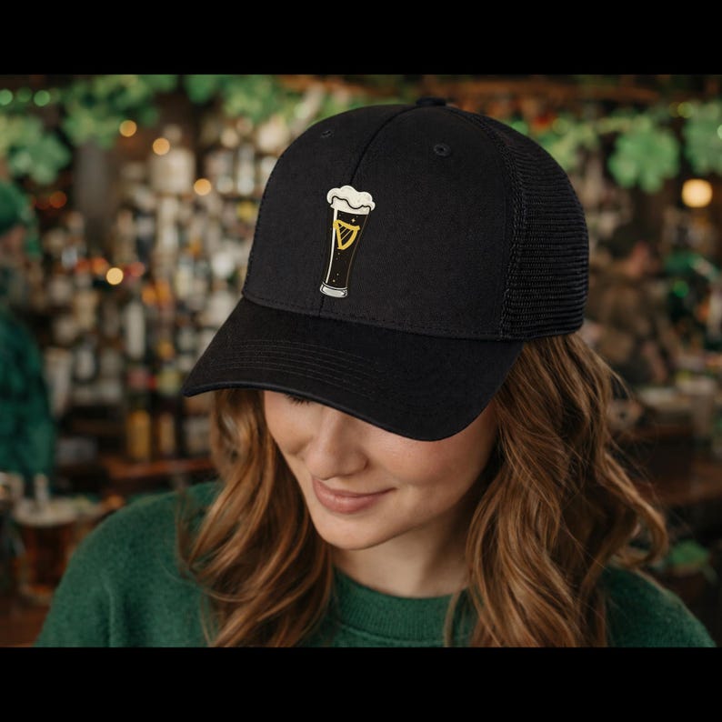 May include: Black baseball cap with a mesh back and a beer glass graphic. The graphic is yellow and white, with a black outline. The cap is worn by a person with long, wavy brown hair.