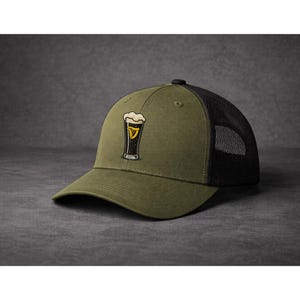 May include: Olive green baseball cap with a black mesh back. The cap features an embroidered design of a pint of stout with a foamy head and a gold harp emblem.