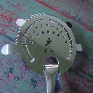 May include: A stainless steel cocktail strainer with a circular perforated head and a handle. The strainer features a small silhouette of a man walking. The handle is curved and the strainer has two small tabs on either side.