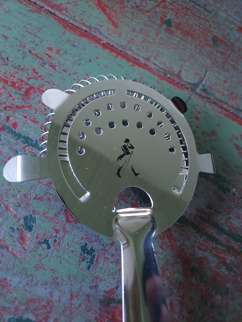 May include: A stainless steel cocktail strainer with a circular perforated head and a handle. The strainer features a small silhouette of a man walking. The handle is curved and the strainer has two small tabs on either side.