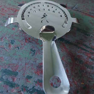 May include: A stainless steel cocktail strainer with a round perforated head and a long handle. The strainer features a small black silhouette of a man walking on the head. The handle has a hole at the end for hanging.