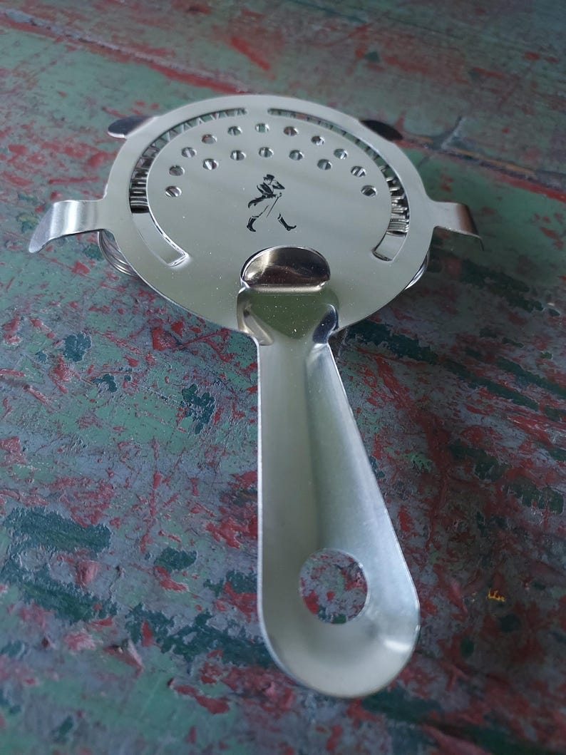 May include: A stainless steel cocktail strainer with a round perforated head and a long handle. The strainer features a small black silhouette of a man walking on the head. The handle has a hole at the end for hanging.