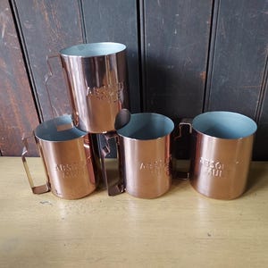 May include: Four copper-colored metal mugs with handles. The mugs have a cylindrical shape with a slightly flared top and a light blue interior. The text "ABSOLUTE MULE" is embossed on the side of each mug. The mugs are arranged on a wooden surface.