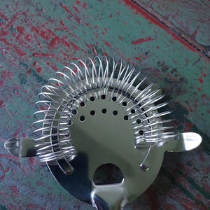 May include: A stainless steel cocktail strainer with a circular head and a handle. The strainer features a coiled spring design around the edge and small holes in the center. The handle has a rounded end.
