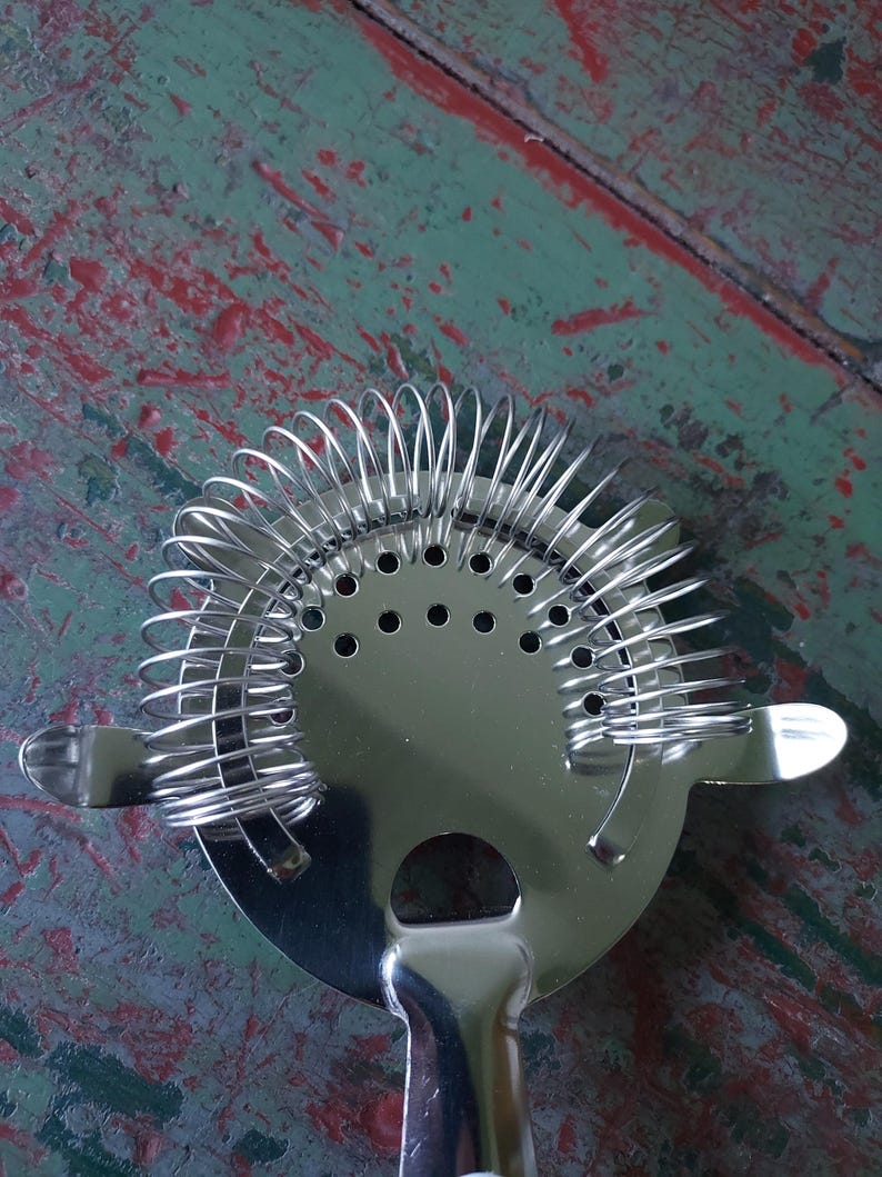 May include: A stainless steel cocktail strainer with a circular head and a handle. The strainer features a coiled spring design around the edge and small holes in the center. The handle has a rounded end.