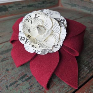 May include: A burgundy felt flower with a layered paper flower center. The paper flower has text printed on it. The felt flower has three large petals and is likely a hair accessory or brooch.
