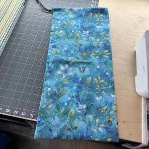 Puede incluir: Una bolsa con cordón con un estampado floral azul. La tela presenta un diseño celestial con hojas y flores azules, verdes y doradas. La bolsa es rectangular y tiene un cierre de cordón.