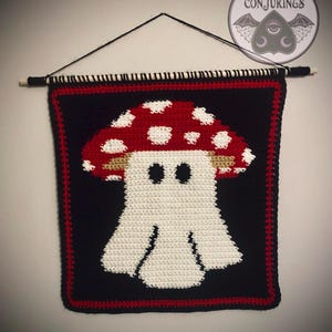 May include: A crochet wall hanging with a mushroom ghost design. The mushroom has a red cap with white spots, a tan stem, and a white ghost body. The background is black with a red border. The text "Cozy Conjurings" is in a circle at the top.
