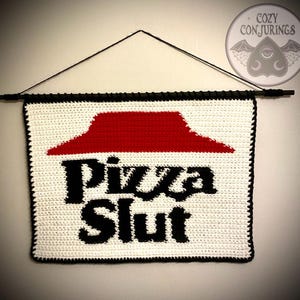 May include: Crocheted wall hanging featuring the words "Pizza Slut" in black, with a red pizza slice logo on a white background. The banner is framed with black yarn and hangs from a black rod.