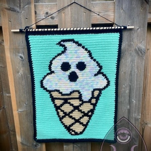 Puede incluir: Un tapiz rectangular de ganchillo con un fondo verde menta claro y un gráfico central de un fantasma blanco con forma de helado. El fantasma tiene ojos y boca negros, delineados con hilo negro sobre un cono de barquillo de color tostado.