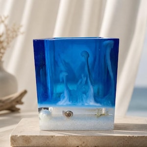 May include: A square resin container featuring a deep blue translucent upper section with suspended white jellyfish-like shapes. The base is clear with embedded small seashells and white sand, creating a layered underwater scene within the object.