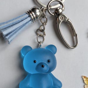 May include: A keyring featuring a translucent blue frosted bear charm with black painted eyes and paw details. The bear is attached to a silver-toned metal split ring and a lobster clasp, accompanied by a light blue suede-like fabric tassel.