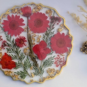 Puede incluir: Un posavasos redondo de resina con borde dorado festoneado, que contiene flores rojas prensadas, follaje verde y copos de pan de oro. El diseño transparente muestra un arreglo botánico de diversas flores rojas y tallos.