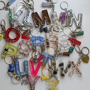Könnte beinhalten: Eine Sammlung von Schlüsselanhängern in Form von Buchstaben aus Kunstharz, die jeweils einzigartige Designs mit Glitzer, Trockenblumen, Tiermustern, eingebetteten Objekten und bunten Mustern aufweisen. Jeder Buchstabe ist an einem Metallring befestigt.
