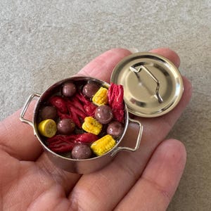 Puede incluir: Olla plateada en miniatura llena de cangrejos de río, maíz y otros ingredientes. La olla tiene dos asas y una tapa. La comida es roja, amarilla y marrón. También se ve una rodaja de limón en miniatura.