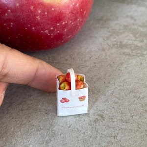 May include: A small white paper bag with a handle, filled with miniature red apples. The bag has a printed design of apples and a pie.