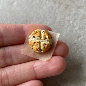 May include: A small, round, brown and white bread roll with a cross pattern on top. The roll is on a piece of parchment paper.