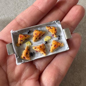 May include: Miniature tray with six golden-brown pastries, each with purple berries, and lemon slices. The pastries sit on a white paper liner inside a silver tray. A few loose berries are scattered around the pastries.