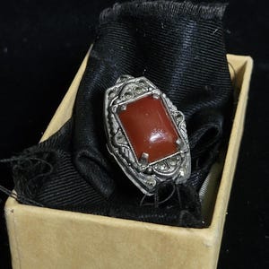 May include: A vintage silver ring featuring a rectangular, reddish-orange gemstone. The ring has an ornate, filigree design and is displayed in a small, beige cardboard box lined with black fabric.