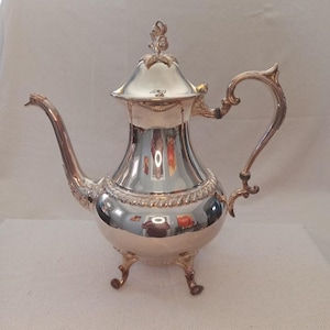 Puede incluir: Una tetera de plata con un asa decorativa y tres patas. La tetera tiene una tapa con un diseño floral.
