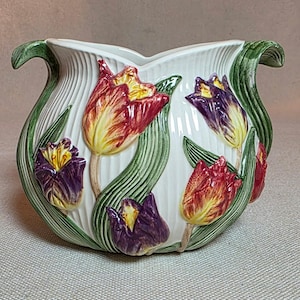 Op de afbeelding: Witte keramische plantenbak met een tulpenontwerp. De plantenbak heeft verticale reliëflijnen en is versierd met rode, gele en paarse tulpenbloemen en groene bladeren. De handvatten zijn groen.